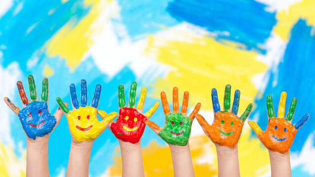 Kids hands covered in vibrant paint with drawn smile icons are raised vertically in a line against an abstract painted background during an art class activity.