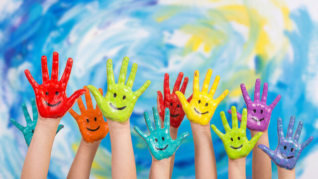 Childrens hands painted in bright multicolored poster paints with smiling face details are held up together in front of a blue and yellow painted artistic background.