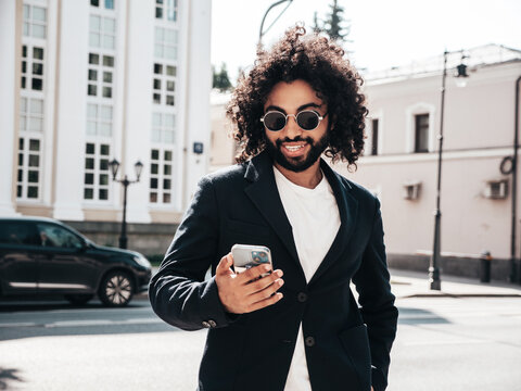 Handsome smiling hipster  model. Sexy unshaven Arabian man dressed in summer suit jacket clothes. Fashion male, long curly hairstyle in the street, using smartphone apps, looking at cellphone screen