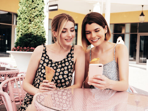 Two young beautiful smiling hipster women in trendy summer dress clothes. Sexy carefree models sitting in terrace cafe. Positive female eating tasty ice cream in waffles cone in sunglasses at sunset