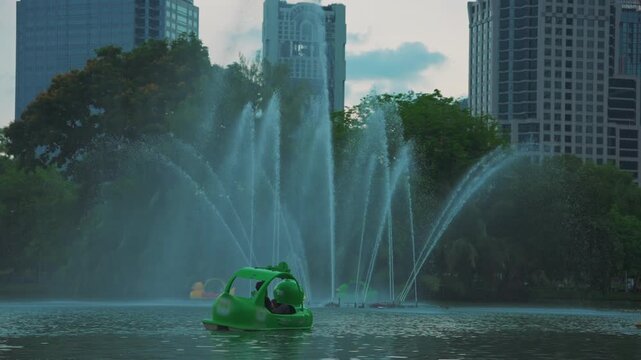 Lumpini Park fountain and pedal boats on lake with Bangkok city skyline, People riding duck boats on water among skyscrapers in Thailand, slow motion 4k