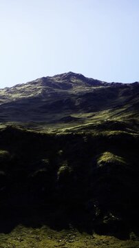 Bright sunlight illuminates the towering mountain, casting long shadows across the rolling hills. Vibrant greenery thrives in the cool air, creating a serene and peaceful landscape scene.