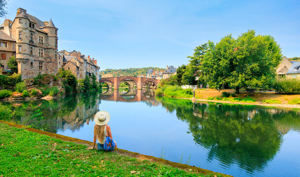 woman sitting on river bank looking at Espalion village and bridge, tour tourism in Aveyron, France