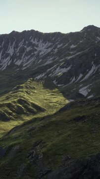 Sunlight gently bathes lush green slopes as shadows play across the rugged mountain landscape. Peaks rise majestically under a clear sky, creating a serene and captivating atmosphere.
