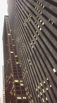 Tall buildings stretch upwards towards the overcast sky as evening approaches. Soft lights glow from the windows, creating a striking contrast against the darkening surroundings.