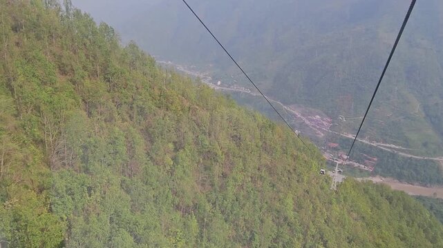 Scenic footage from the Manakamana cable car showing a passenger gondola traveling above densely forested mountain slopes, winding river valley, roads, settlements, and surrounding Himalayan foothills