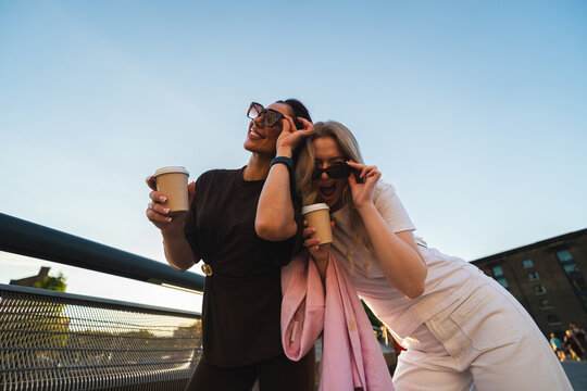 Two cheerful female friends holding coffee cups and smiling on an urban bridge