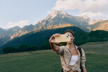 Naklejka premium A woman in casual attire takes a selfie outdoors in a green field, with mountains and blue sky behind, highlighting lifestyle moments in nature and travel.