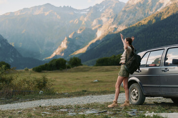 Naklejka premium Woman with backpack standing near car on mountain road pointing at distant peaks. Outdoor lifestyle, adventure, travel, and nature exploration concept in warm daylight.