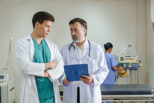 Caucasian senior doctor and asian young doctor discussing emergency care details in hospital examination room while nurse working with equipment in background during real time coordination