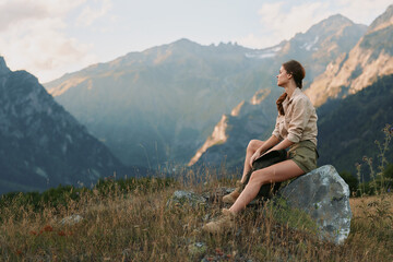 Fototapeta premium A woman in casual clothes sits on a rock in a mountain meadow, looking at the distant peaks during a calm sunset. Concept for lifestyle, nature, and relaxation.