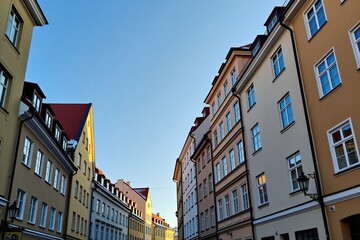 Obraz premium Low angle view of narrow city street with tall historical buildings on clear day. Sunlight illuminates colorful house facades under blue sky. Old town architecture in Europe stands along alleyway.