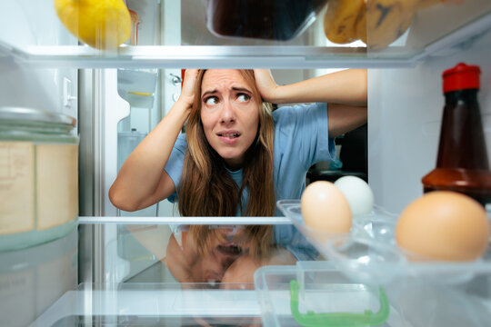 Shocked Woman Looking In Refrigerator Finding Nearly Empty Shelves In The Morning. Upset lady holds her head in shock looking inside a nearly empty fridge