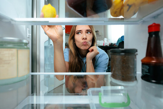 Woman Choosing Fresh Lemon From Refrigerator Healthy Breakfast In The Morning. Uncertain lady reaches for a fresh lemon inside an open kitchen fridge