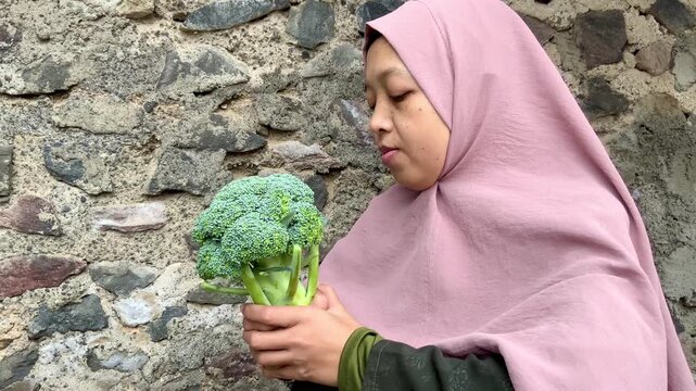Woman wearing a hijab holding a fresh head of broccoli against a stone wall background