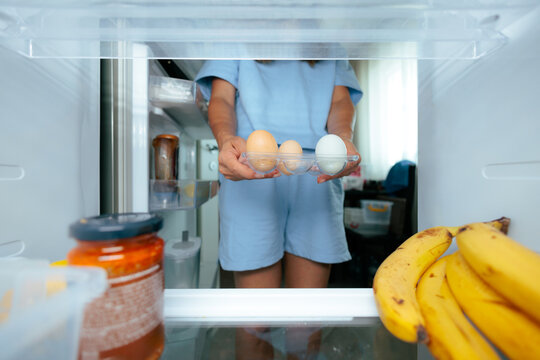 Woman Holding Small Egg Tray Inside Refrigerator In The Morning. Person holds a transparent plastic tray with eggs inside an open kitchen fridge in the morning.
