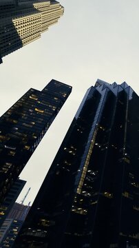 Tall, striking skyscrapers reach towards a gray sky, their windows aglow with warm light as evening settles in. The architecture showcases modern design and urban beauty.