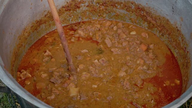 A large cauldron is used to prepare food at a street festival. People gather around to watch as the dish simmers and the aroma fills the area.