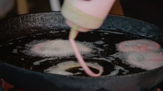 A cook prepares colorful jalebi by squeezing batter into hot oil at a busy street food festival. The oil bubbles around the fried dessert, attracting many onlookers.