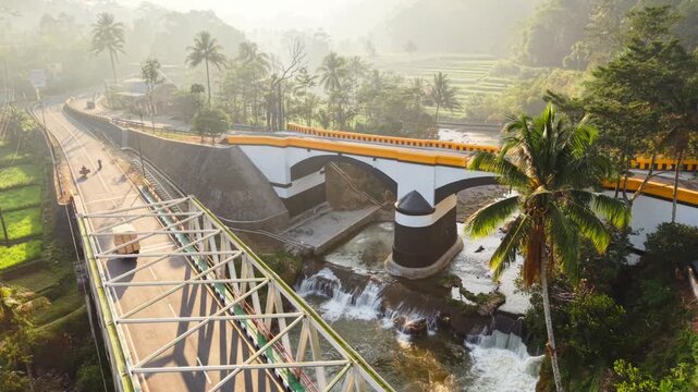 Aerial view of a metal truss bridge and an arched bridge crossing a river in a lush tropical landscape with palm trees and rice fields in Indonesia.