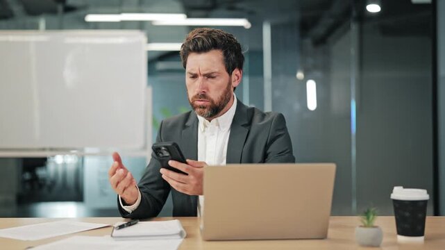 Businessman expressing frustration while using smartphone. Displays worry confusion. Faced with a problem or unexpected situation at office desk, reflecting technology challenges and business stress.