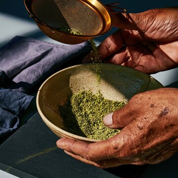 Hands of an older East Asian man sifting green matcha powder into a crackled raku bowl, weathered skin and tactile textures for craft and tradition