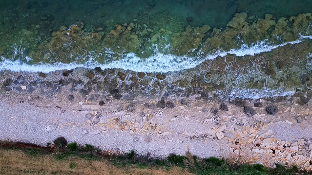 Aerial view captures a rugged coastline with crashing waves against rocky shores. The beach is a mix of pebbles and sand, bordered by patches of grass, showcasing raw, natural seaside beauty.