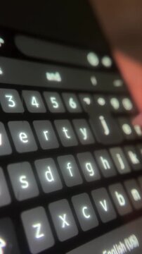 Close up of a finger texting on a smartphone's digital keyboard, scrolling through social media, and chatting on a mobile device in a dark room illuminated by the screen