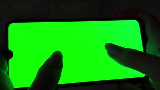 Woman's hand holding smartphone and using gestures like touching, swiping and scrolling on a green screen display in a dark room. Blank chroma key for custom content