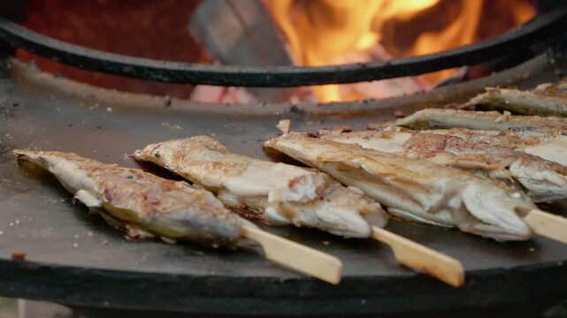 Trout is grilling on sticks over an open fire at a lively street food festival. The aroma fills the air as people gather to enjoy the tasty dish.
