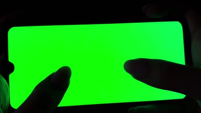 Woman's hands using a smartphone with a green screen in a dark room, scrolling through social media, swiping pages, and tapping on the touchscreen display for interactive use
