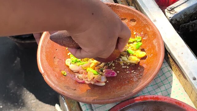 Grinding fresh chili ingredients in a clay mortar. A close-up shot of a hand using a wooden pestle to grind fresh shallots, chili peppers, and garlic in a traditional clay mortar. 4K 60fps food video.