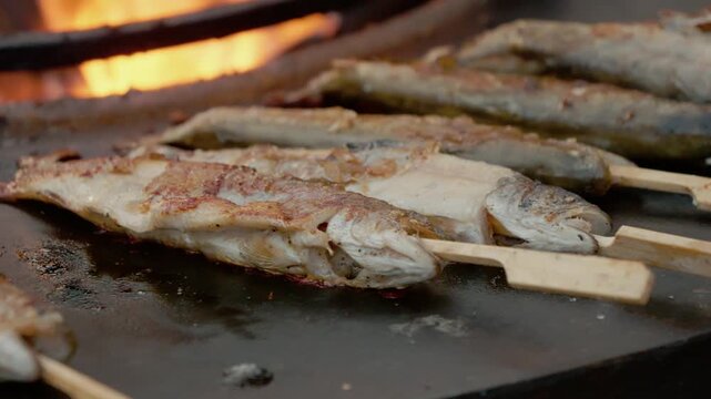 Trout cooks on skewers over an open flame at a lively street food festival. The food attracts many people enjoying the warm evening atmosphere.