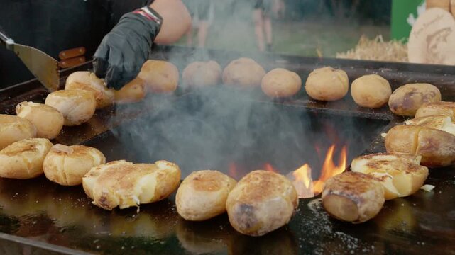 Whole baked potatoes are cooking on a grill plate with a fire at a street food festival. The sound of sizzling fills the air as people gather around to enjoy.