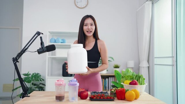 Sporty Asian woman broadcasting vegan lifestyle tips using mobile phone on tripod. Energetic health content creator sharing raw food recipes and fitness motivation online.