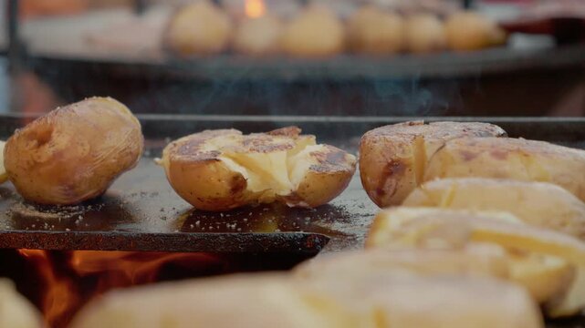 A group of whole baked potatoes sits on a grill plate with flames below. The potatoes are cooking at a street food festival, creating a delicious aroma.