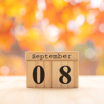 Natural wooden cubes with text SEPTEMBER 8, minimalist calendar reminder on wooden surface, soft orange autumn leaves blurred on background, warm seasonal tones. Close-up.