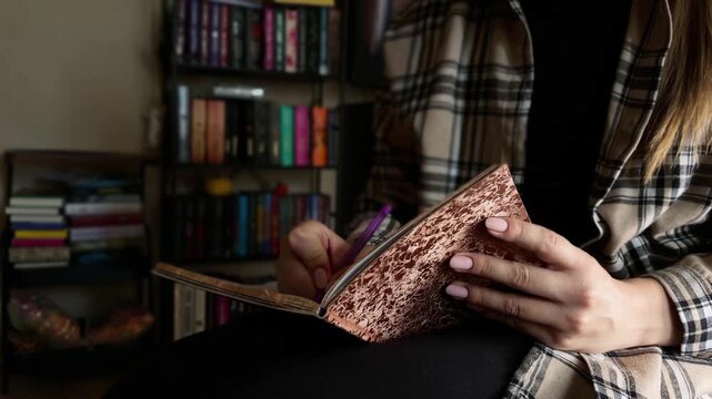 Woman's hands writing notes in a diary journal with a pen. Unrecognizable female taking notes in a notebook while sitting in a library next to a bookshelf at home