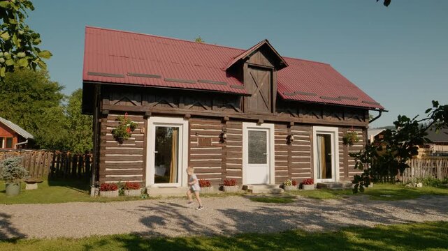 A boy walks outside a traditional house in Bucovina. He pauses and then looks at the camera. The house has wooden walls and flowers outside.