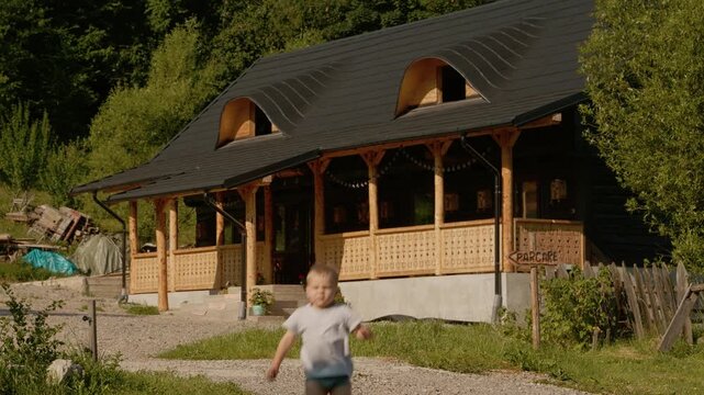A little boy runs across the yard toward a traditional house in Bucovina. The house is surrounded by greenery. Flowers and plants are visible near the entrance.