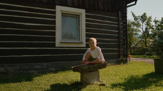 A boy looks at the camera while seated on a stone in Bucovina. The traditional house stands behind him, showing the style of the region.