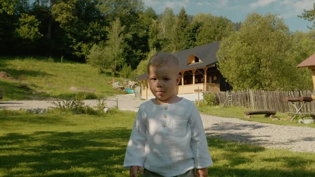 A boy stands on green grass and looks at the camera. Behind him is a traditional house in Bucovina. Trees surround the area under a clear sky.