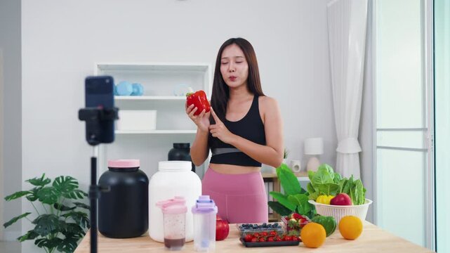 Female fitness coach filming post-workout nutrition guide with fresh green veggies. Smiling Asian girl recording healthy smoothie preparation for wellness social media channel.