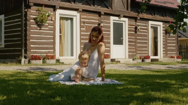 A mother sits on the grass with her child in front of a traditional house in Bucovina. They spend time together, enjoying the warm sun and the outdoors.