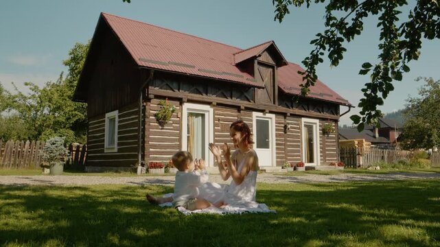A mother and child sit on the grass and play together in front of a traditional house in Bucovina. They enjoy their time under the sun surrounded by nature.