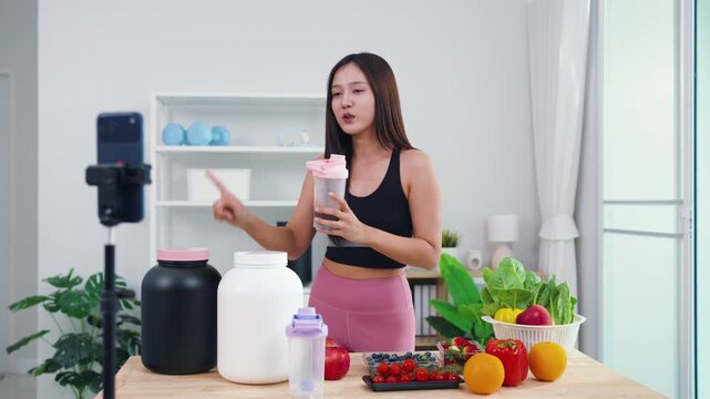 Female fitness coach filming post-workout nutrition guide with fresh green veggies. Smiling Asian girl recording healthy smoothie preparation for wellness social media channel.
