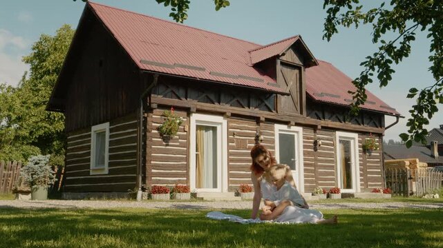A mother sits with her child on the grass, reading a book. They are in front of a traditional house in Bucovina. The sun lights up the scene and nearby plants.