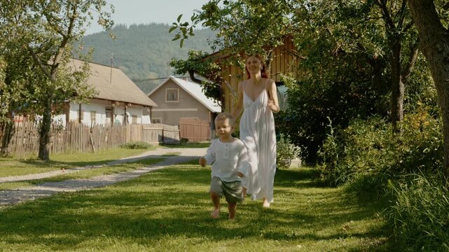 A woman and her boy play together as they run through a yard. This takes place in Bucovina near traditional houses surrounded by mountains and greenery.