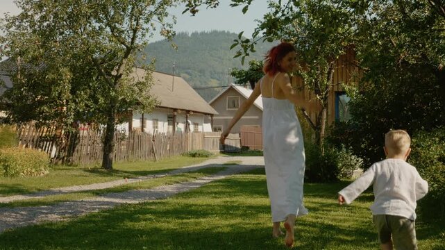 A woman and her young boy run through a yard in the mountains of Bucovina. They enjoy their time outside among the trees and the house.