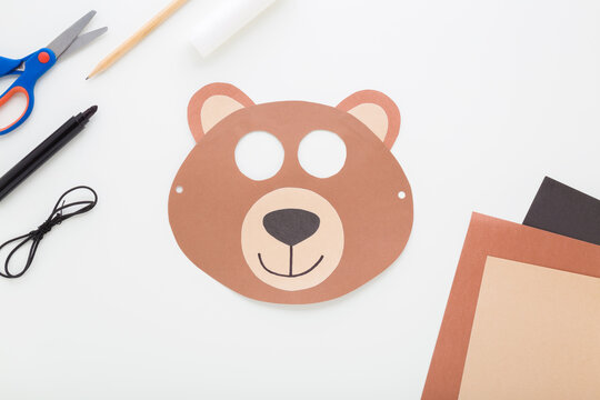 Brown smiling bear mask with scissors, glue, pencil, marker, string and application paper on light gray table background. Closeup. Creating decoration for kid. Flat lay. Top down view.